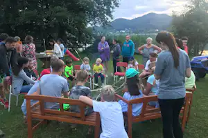 Familie sitzt im Kreis auf Holzstühlen im Campinggarten, im Hintergrund Bäume, See und Berge.