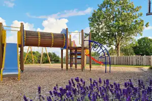 Spielplatz mit Rutsche und Klettergerüst auf Kies, davor Lavendelblumen und ein Baum am Campingplatz.