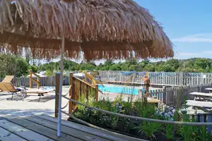 Holzliege unter Strohdach am Campingplatz, dahinter ein Pool und ein Holzsteg mit Pflanzenbeeten und Zäunen.