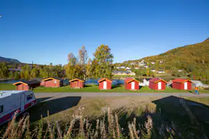 Campingplatz mit roten Hütten, Rasenfläche und Wegen, im Hintergrund Bäume und ein bewaldeter Hügel.