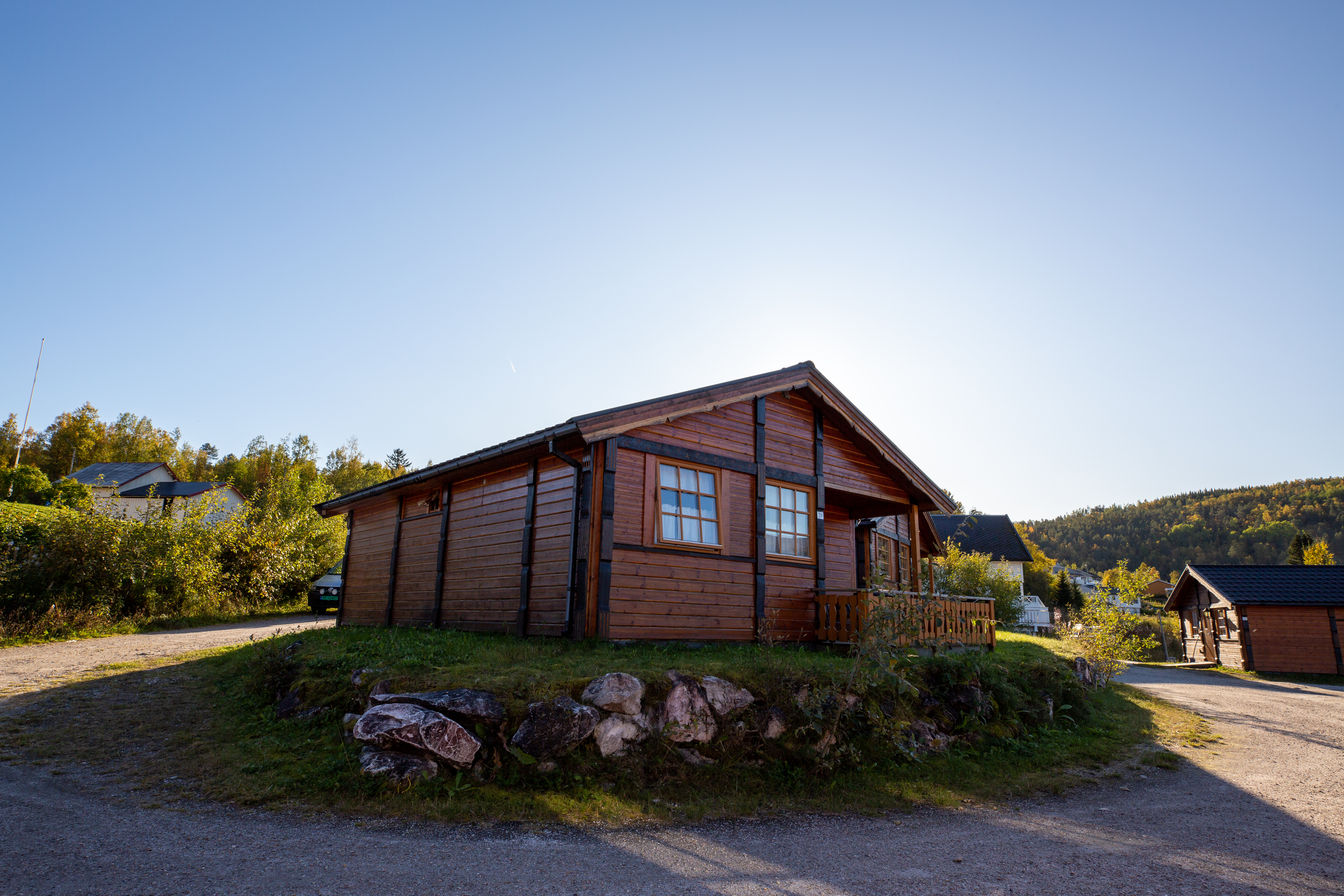 Holzhütte auf einem Campingplatz mit Kiesweg, kleinen Büschen und Rasenfläche, im Hintergrund weitere Gebäude.