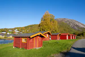 Rote Holzunterkünfte am Flussufer, mit Grasfläche, Weg und weiteren Gebäuden im Hintergrund.