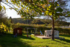 Spielbereich im Campinggarten mit Sandkasten, Rutsche und Schaukel vor einem kleinen Holzhaus, umgeben von Bäumen und Wiese.