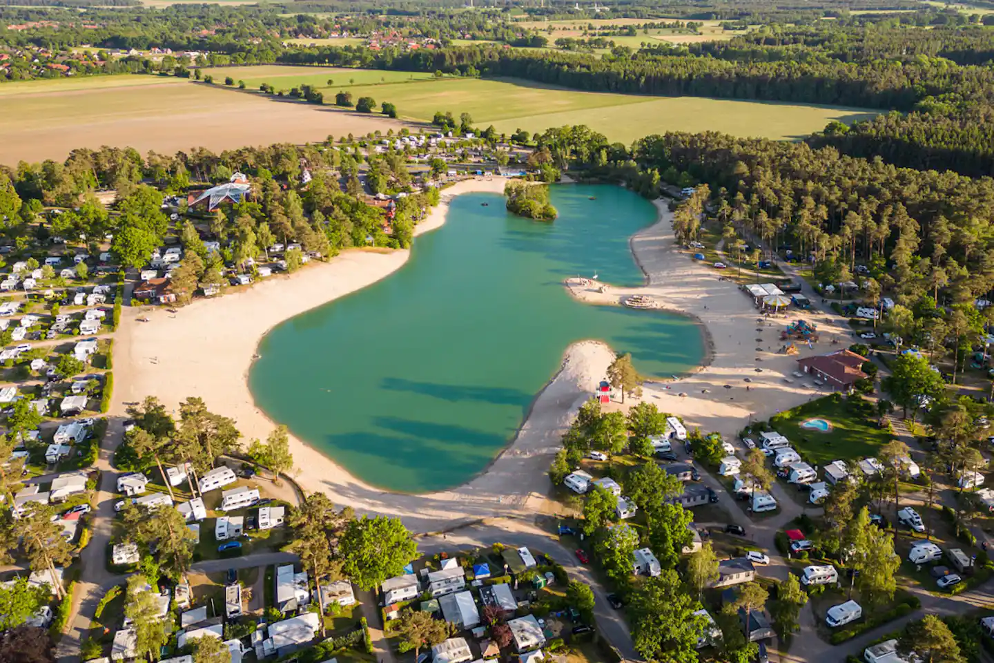 Luftaufnahme vom Campingplatz am See mit türkisfarbenem Wasser, Sandstrand, Booten und Stellplätzen zwischen Bäumen.