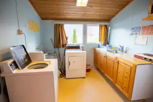 Innenansicht eines Wohnwagens mit Küchenzeile, Spüle, Herd und Waschmaschine, daneben Fenster mit Vorhängen.