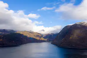 Blick auf einen See zwischen Bergen, umgeben von bewachsenen Hängen und großen Wolken am Himmel.