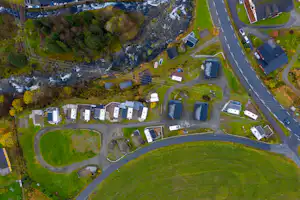 Luftaufnahme einer Campinganlage mit mehreren Gebäuden, Stellplätzen, Zufahrtsstraße und einem Fluss mit Stromschnellen.