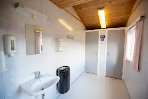 Sauberes Sanitärgebäude mit Waschbecken, Mülleimer und WC-Tür, Holzdecke und Fliesenboden im Innenraum.