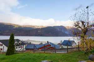 Blick auf Dächer von Ferienhäusern am See, im Hintergrund Berge und Wolken, rechts ein Baum mit gelben Blättern.