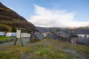 Campingplatz mit Wohnwagen und Stellplätzen, Steinterrasse mit Holzgeländer, Blick ins Tal und auf bewaldete Berge.