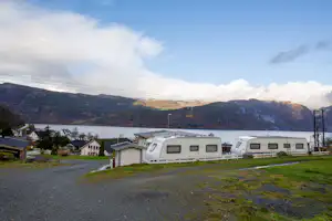 Wohnwagen auf einem Campingplatz am See, im Hintergrund Berge und Häuser am Ufer.