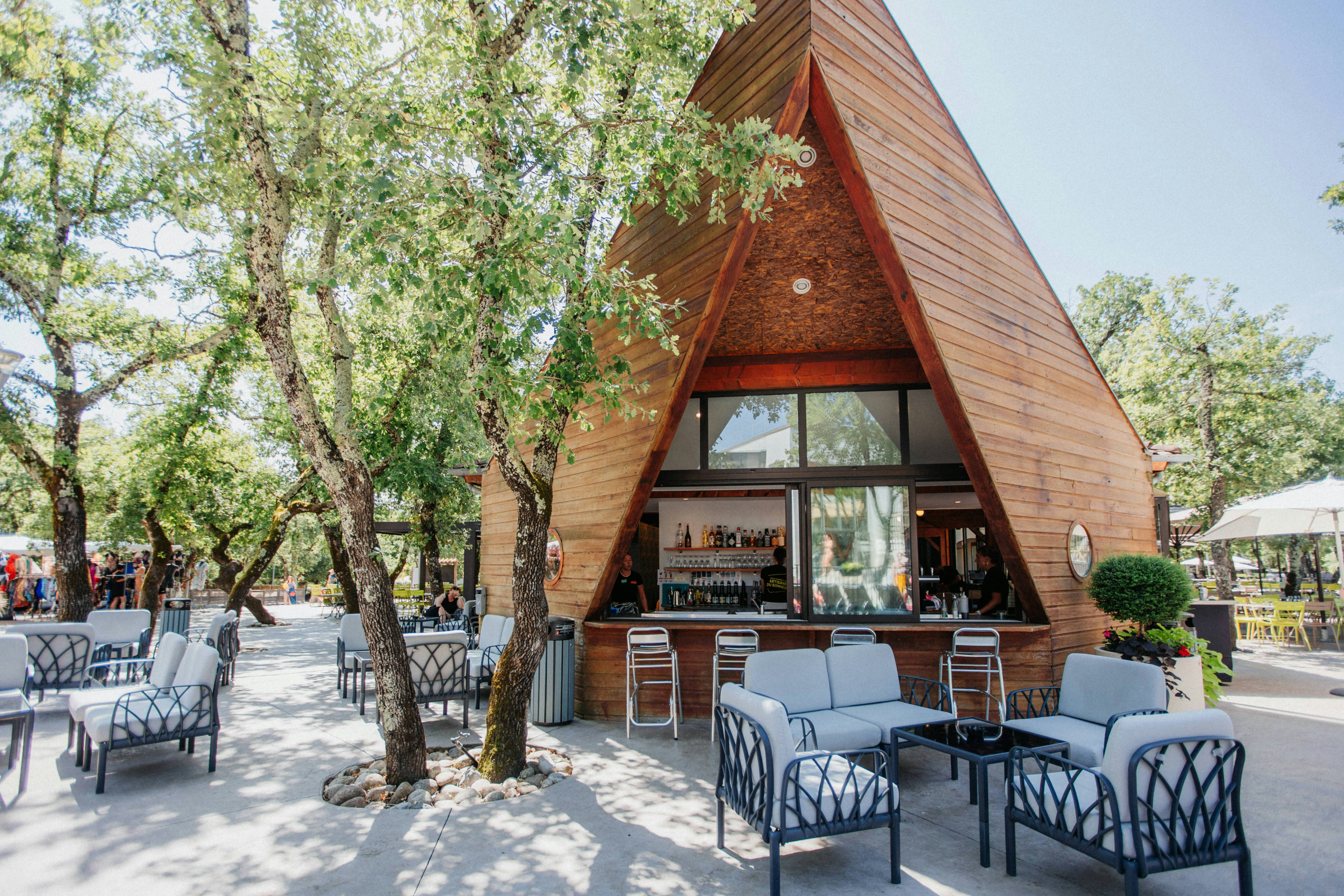 Holzhaus mit großen Fenstern und Terrasse, davor Gartenmöbel und Sonnenschirme, umgeben von Bäumen im Campingbereich.