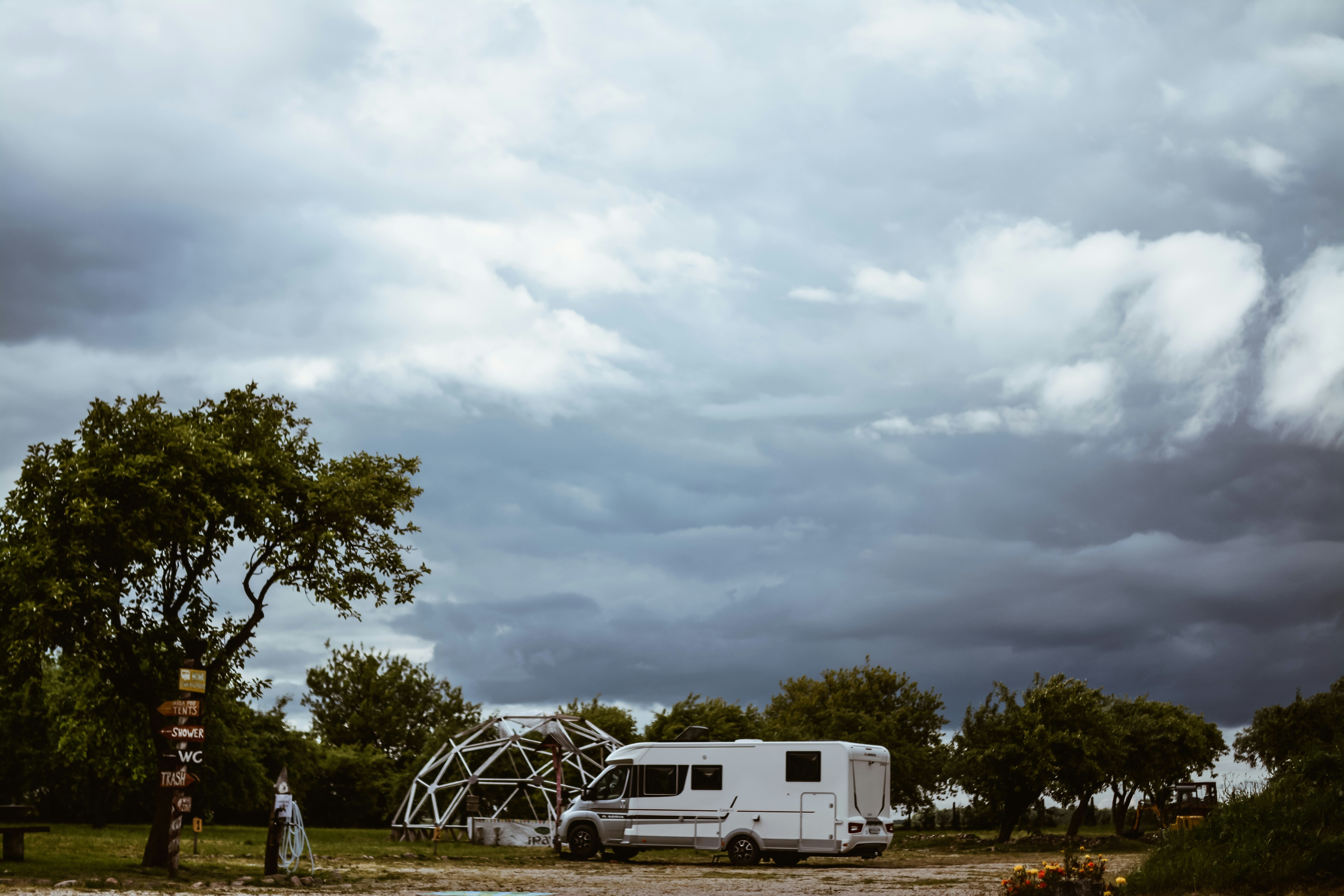 Wohnmobil vor einem geodätischen Dome auf dem Campingplatz, umgeben von Bäumen und dunklen Wolken am Himmel.