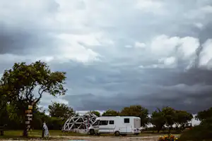 Wohnmobil vor einem geodätischen Dome auf dem Campingplatz, umgeben von Bäumen und dunklen Wolken am Himmel.