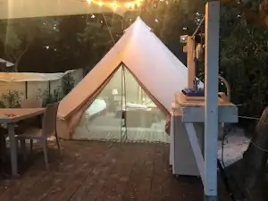 Weißes Zelt auf einer Holzterrasse mit Glasfront und Sitzbereich, im Hintergrund weitere Zelte und Bäume.
