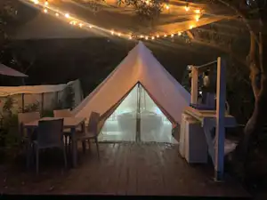 Beleuchtetes Campingzelt auf Holzterrasse mit Lichterkette, Tisch und Stühlen am Rand, im Hintergrund Bäume.