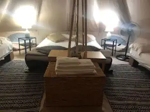 Innenansicht eines Zelts mit Doppelbett, Nachttischen, Tisch und Ventilator auf Teppichboden.
