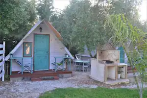 Holz-Campinghütte mit blauer Tür und Veranda, daneben Sitzgruppe und Grillstelle im Garten mit Bäumen und Gras.