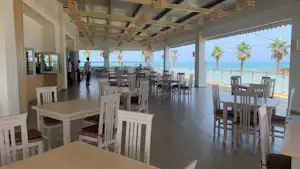 Restaurantbereich auf einer Camping-Terrasse mit Holzdach, vielen Tischen und Stühlen sowie Blick auf das Meer und Palmen.