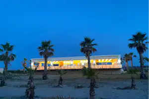 Hotelgebäude am Campingplatz mit beleuchteter Fassade, davor mehrere Palmen im Sand bei blauem Abendhimmel.