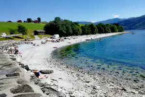 Badeufer am See mit Kiesstrand, Campingzelten und Wohnwagen am Rand, im Hintergrund Berge und grünes Ufer.