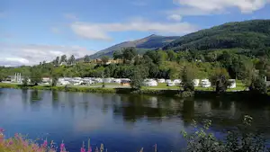 See mit spiegelndem Wasser, am Ufer stehen Wohnwagen und Campingfahrzeuge vor grünen Hügeln und Bäumen.