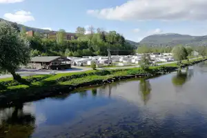 Campingplatz am Fluss mit Wohnwagen und Stellplätzen, gepflegtem Grün, Häusern im Tal und Bergen im Hintergrund.
