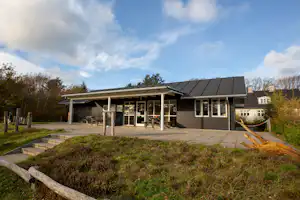 Campinghaus mit überdachter Veranda und Terrasse, gepflasterter Fläche, Bäumen und Rasen im Vordergrund.