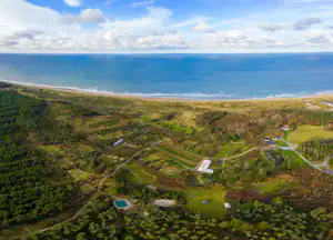 Luftaufnahme einer Küstenlandschaft mit Strand, Wäldern und einem Campingplatz mit Wegen und kleinen Gebäuden.