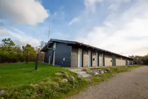 Reihen von grauen Sanitärgebäuden am Campingplatz, mit Betonweg, Rasenfläche und Bäumen am Rand.