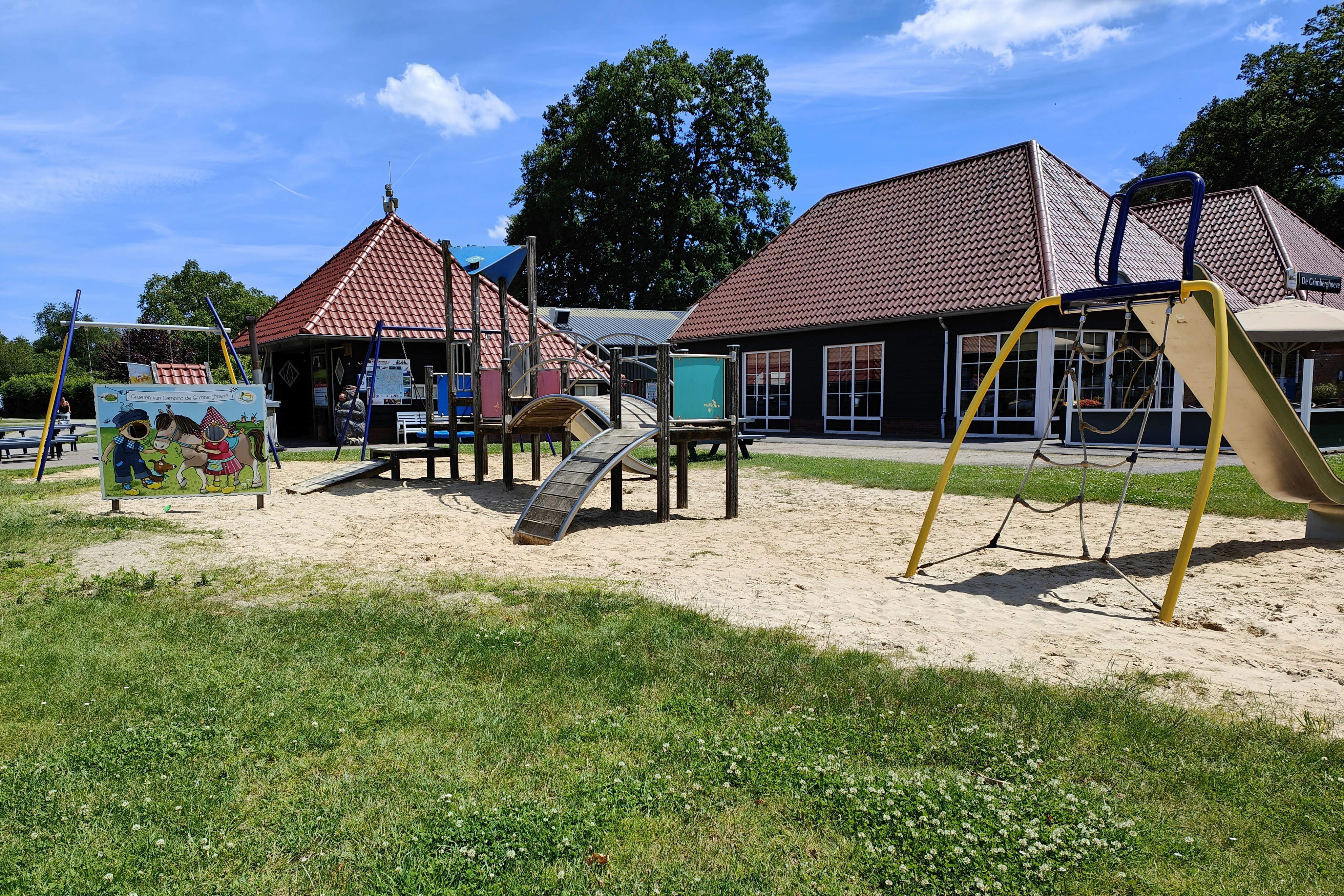 Spielplatz auf dem Campingplatz mit gelber Schaukel, Rutsche, Sandfläche und Holzhäusern mit roten Ziegeldächern.