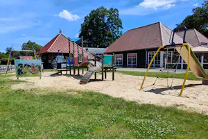 Spielplatz auf dem Campingplatz mit gelber Schaukel, Rutsche, Sandfläche und Holzhäusern mit roten Ziegeldächern.