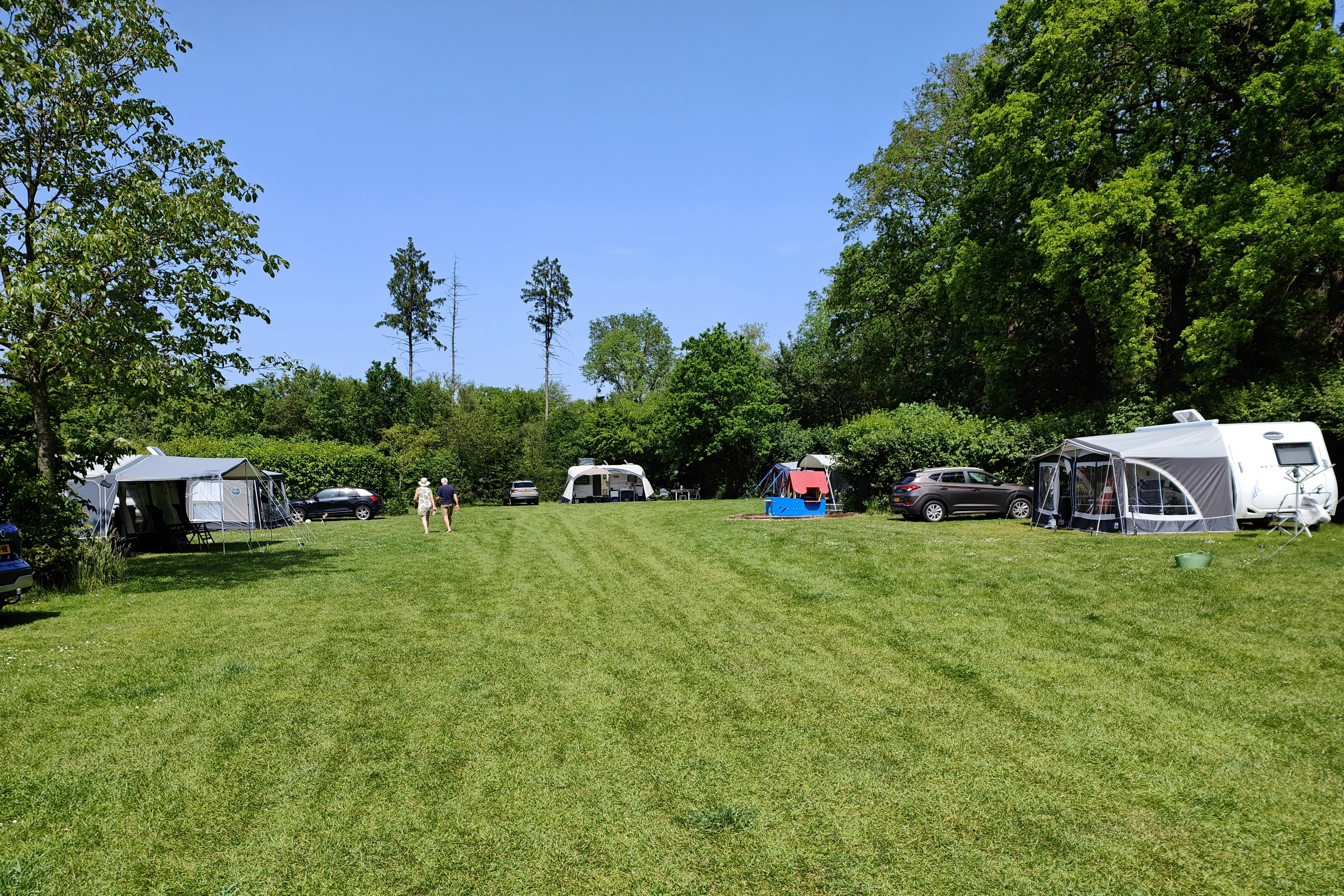 Campingplatz mit Wohnwagen und Zelten, Autos auf der Wiese, Bäume am Rand und ein großes Zelt rechts.