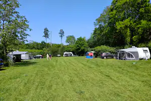 Campingplatz mit Wohnwagen und Zelten, Autos auf der Wiese, Bäume am Rand und ein großes Zelt rechts.
