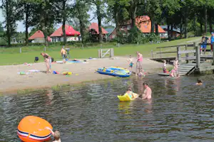 Badebereich am See: Kinder im Wasser, Schwimmreifen und aufblasbares Boot am Sandstrand, Holzsteg und Bäume im Hintergrund.