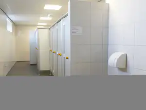 Flur in einem Sanitärgebäude mit Toiletten, weißen Wänden und Türen, daneben ein Spender an der Wand.