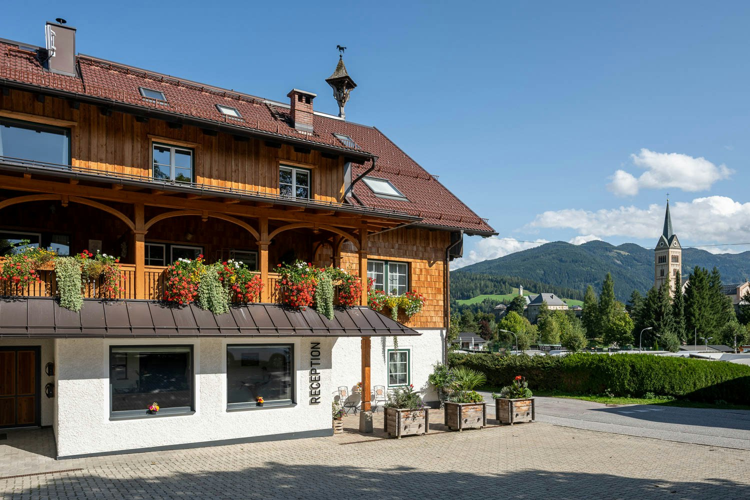 Campinggebäude mit Rezeption, Holzgalerie und roten Dachziegeln, davor Sitzgelegenheiten und Pflanzen auf dem gepflasterten Platz.