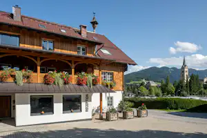 Campinggebäude mit Rezeption, Holzgalerie und roten Dachziegeln, davor Sitzgelegenheiten und Pflanzen auf dem gepflasterten Platz.