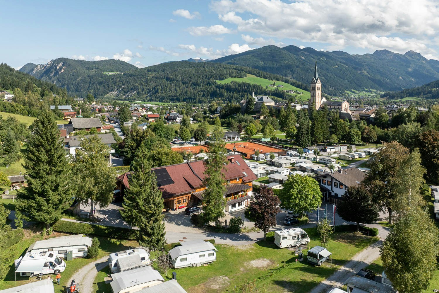 Campingplatz mit Wohnwagen und Stellplätzen, Blick auf ein Dorf mit Kirche und Berge im Hintergrund.