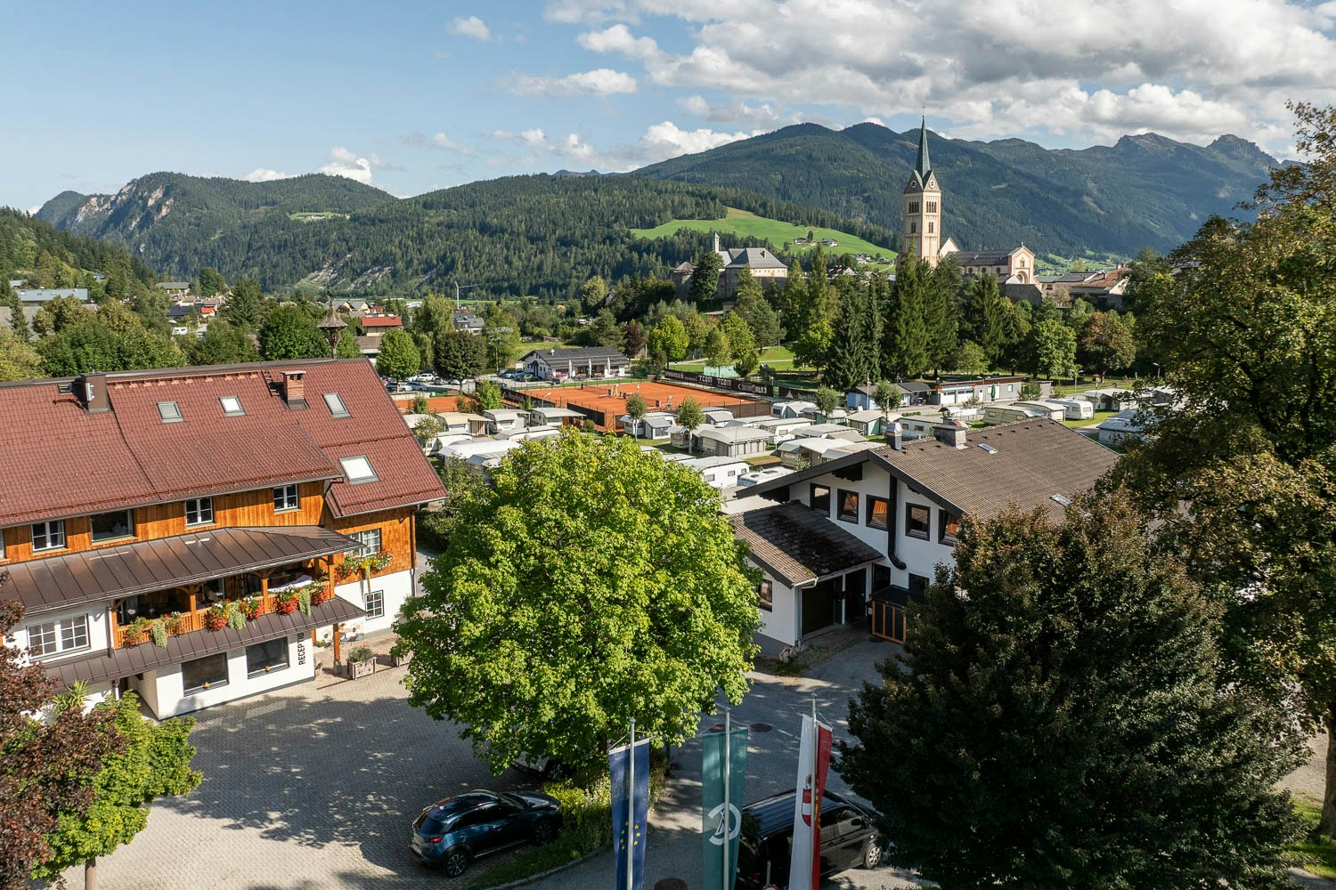 Blick auf ein Campinggelände mit Wohngebäuden, Stellplätzen und Bäumen, im Hintergrund Berge und ein Kirchturm.