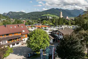 Blick auf ein Campinggelände mit Wohngebäuden, Stellplätzen und Bäumen, im Hintergrund Berge und ein Kirchturm.