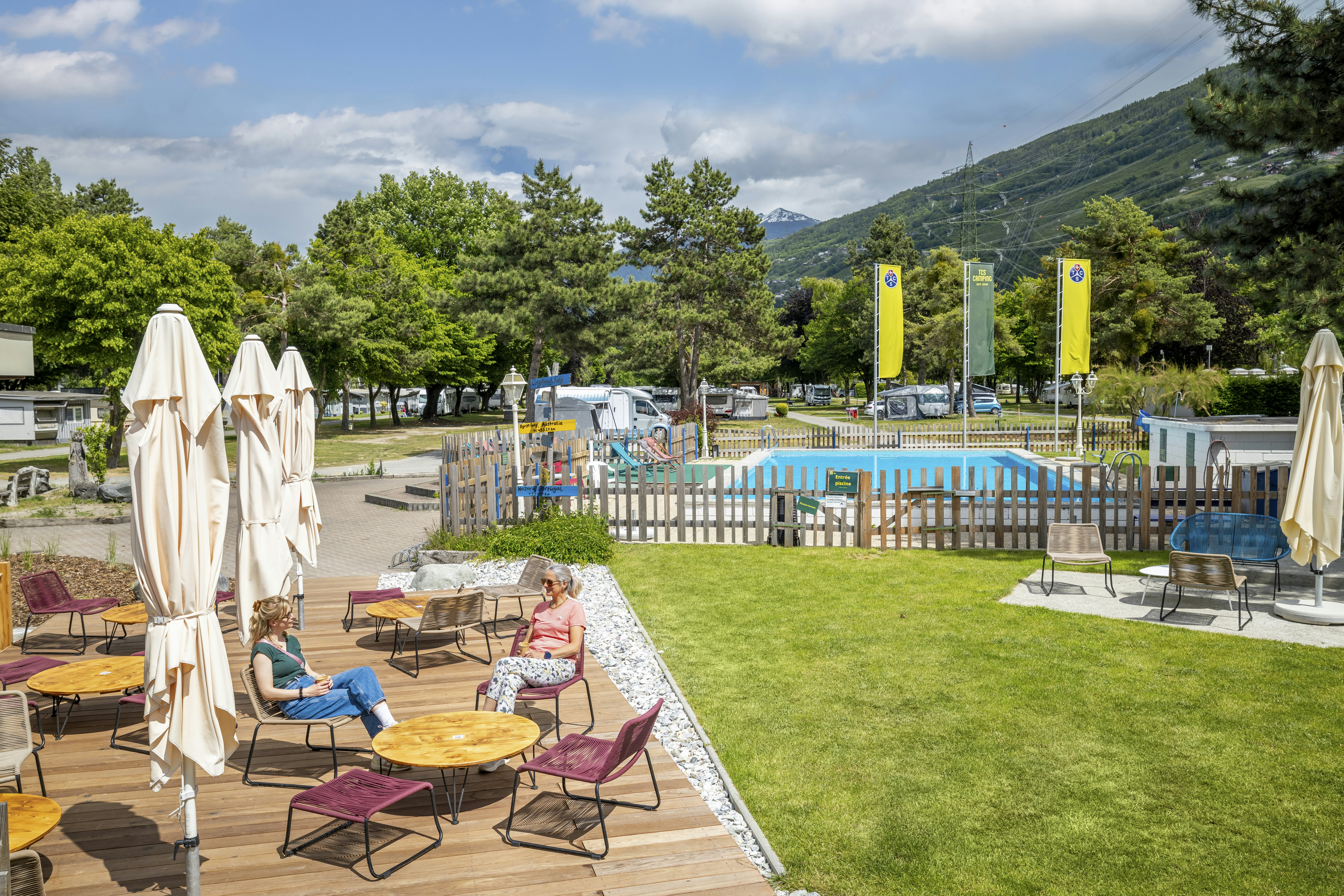 Campingplatz mit Rasenfläche, Sonnenliegen und Tischen, daneben ein kleiner Pool und Bäume sowie Berge im Hintergrund.
