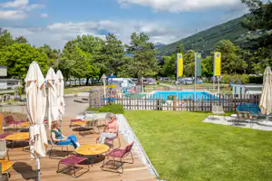 Campingplatz mit Rasenfläche, Sonnenliegen und Tischen, daneben ein kleiner Pool und Bäume sowie Berge im Hintergrund.