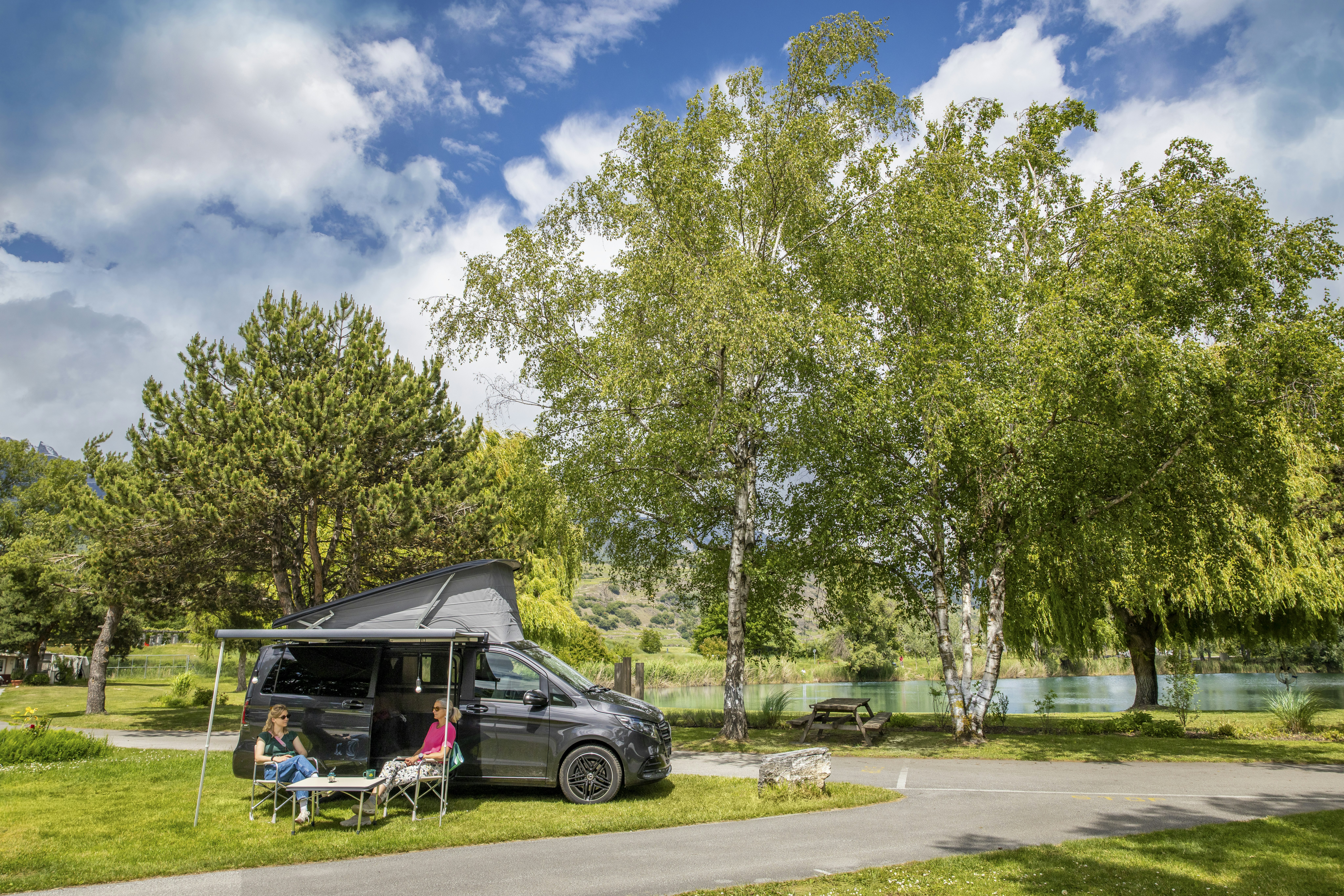 Wohnmobil mit Markise am Stellplatz, davor Campingstühle und Tisch, Bäume und Rasen neben einem Weg.