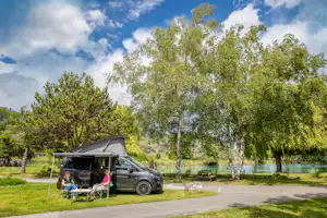 Wohnmobil mit Markise am Stellplatz, davor Campingstühle und Tisch, Bäume und Rasen neben einem Weg.