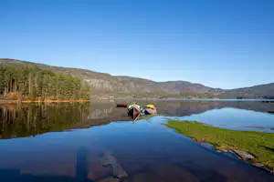 Ruderboot am Steg am Seeufer, umgeben von Waldhängen und spiegelndem Wasser unter klarem Himmel.