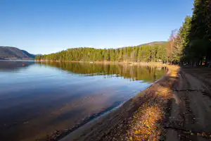 Ruhiger See mit Uferweg, Wald und Bergen in der Ferne, spiegelnde Wasseroberfläche und Bäume am Rand.
