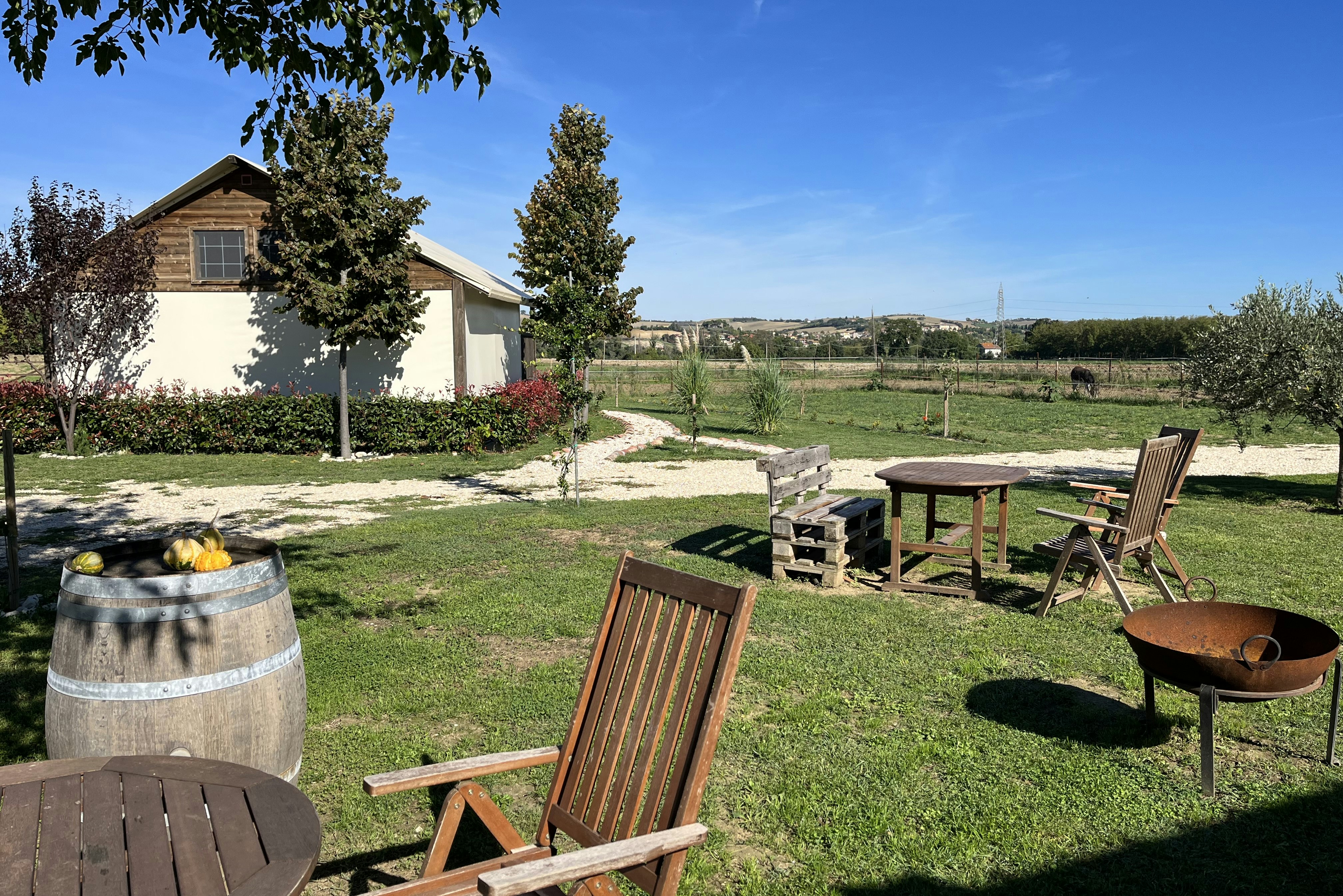 Holzhütte auf dem Campingplatz mit Sitzgruppe, Feuerstelle und Gartenbeeten, im Hintergrund Wiese und Bäume.