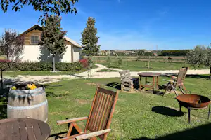 Holzhütte auf dem Campingplatz mit Sitzgruppe, Feuerstelle und Gartenbeeten, im Hintergrund Wiese und Bäume.