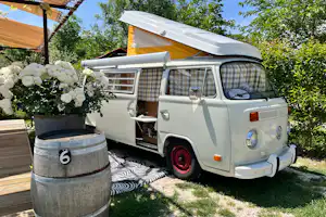 Weißer Wohnwagen auf dem Campingplatz mit Markise, davor ein Tisch und Blumen in einem Holzfass.